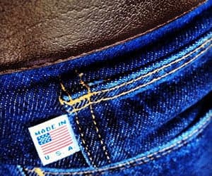 Close-up of denim jeans featuring a small American flag tag near the coin pocket, from All American Clothing Co. promotional imagery.