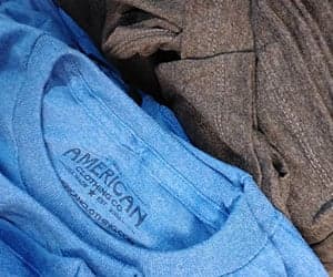 Close-up of folded t-shirts with an All American Clothing Co. neck label, from All American Clothing Co. promotional imagery.